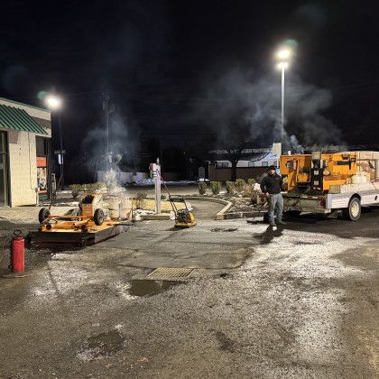 Asphalt paving at night: Workers operate machinery in a parking lot, illuminated by streetlights, with smoke rising.