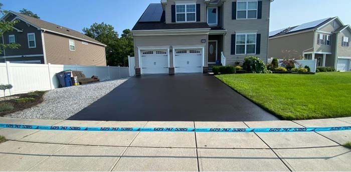 Newly paved black driveway in front of a two-story house with green lawn and blue tape on the sidewalk.