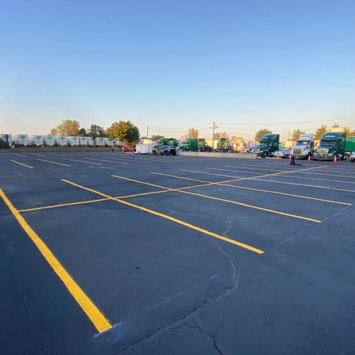 Large asphalt parking lot with yellow lines. Semitrucks and trailers are parked in the distance. Clear blue sky.