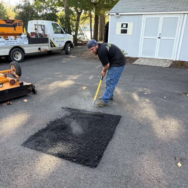 Man raking asphalt patch on driveway, with truck and shed in background.