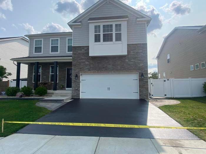 Two-story house with a newly paved black driveway. The house has gray siding and a stone facade.
