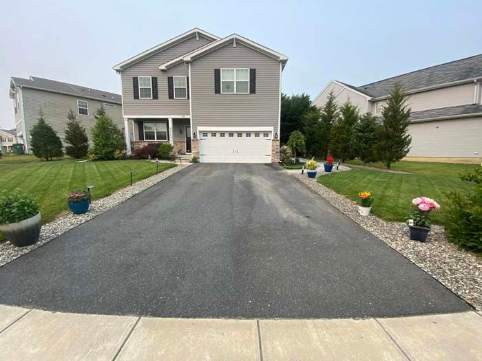 Two-story gray house with a black paved driveway lined with potted plants, lawn on both sides, and cloudy sky.