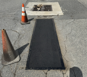 Asphalt patch in street with two traffic cones near a storm drain.