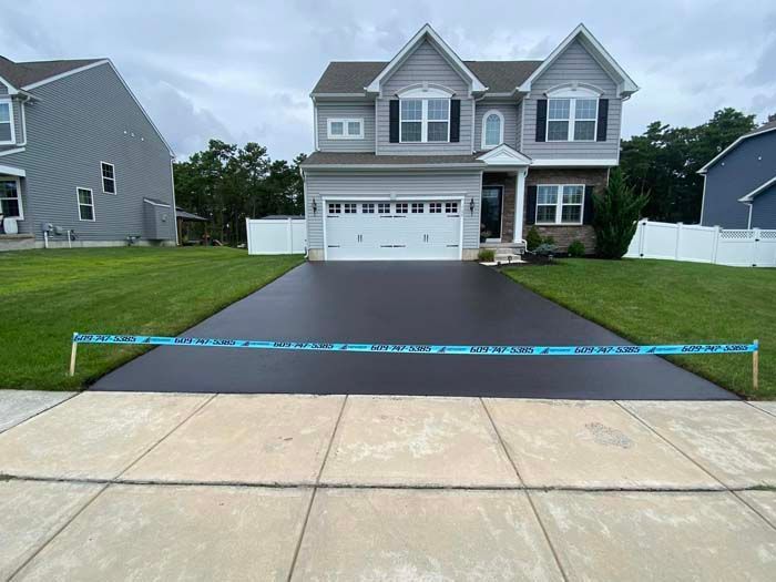 A newly paved black driveway in front of a two-story gray house with green grass and white fence.
