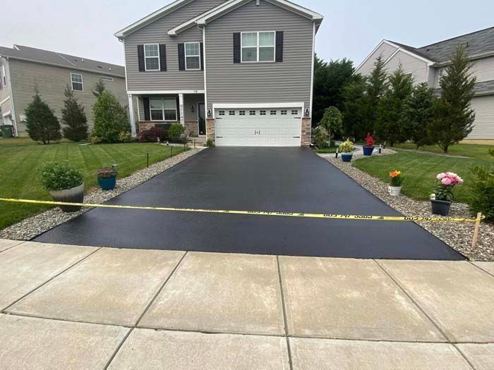 Newly paved asphalt driveway in front of a gray house, bordered by stone and flowers.