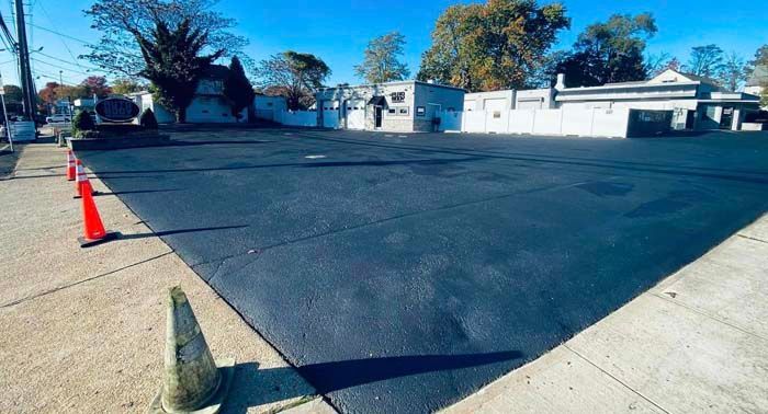 Newly paved asphalt lot with orange cones on the edge, buildings in the background.