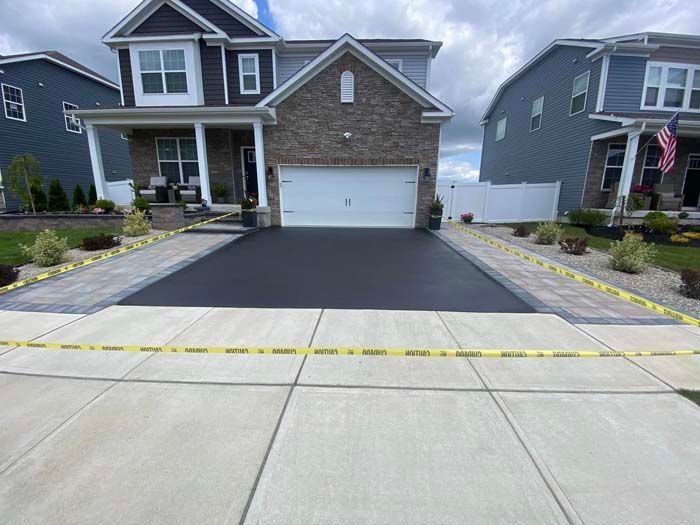 Newly paved black driveway with brick borders, fronting a two-story house. Yellow caution tape is across the driveway.