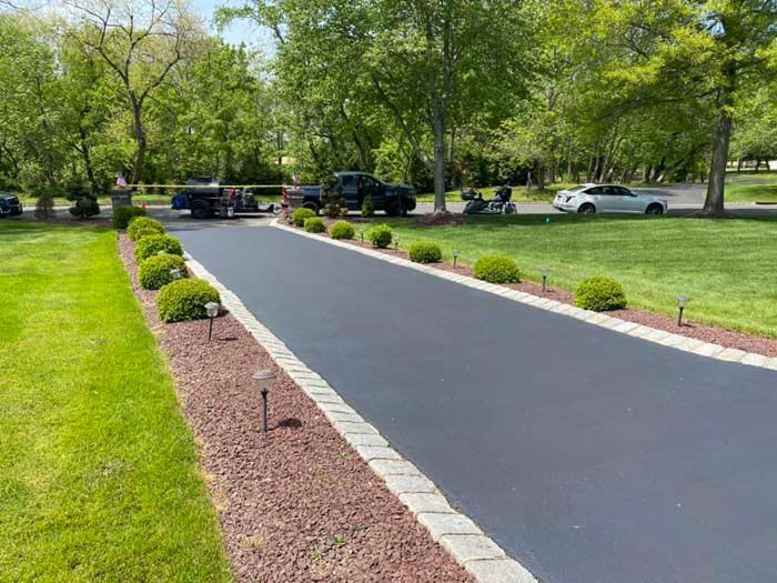 Newly paved asphalt driveway lined with bushes, ground cover, and stone edging; vehicles present.