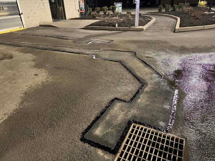 Asphalt area with freshly sealed cracks. A metal grate and curb border the dark pavement.