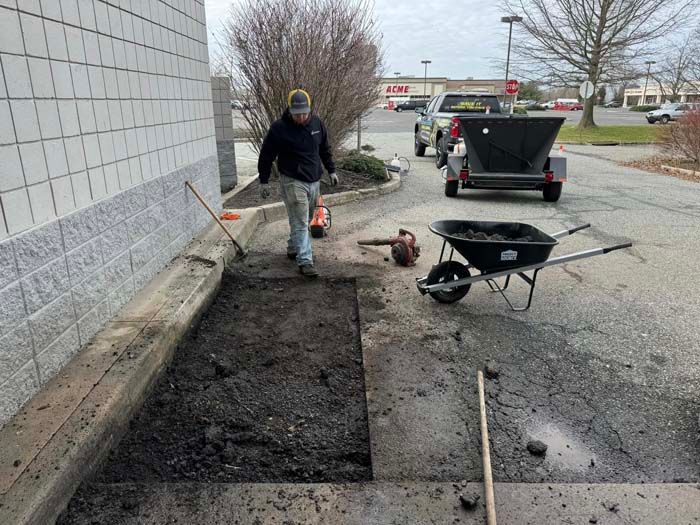 Man repairing asphalt next to a building. A wheelbarrow, tools, and a trailer are nearby.
