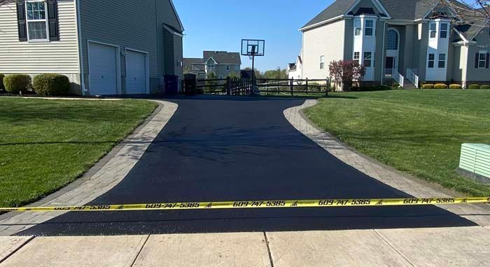 Newly paved black asphalt driveway with yellow caution tape, flanked by green grass and concrete borders, leading to a garage.