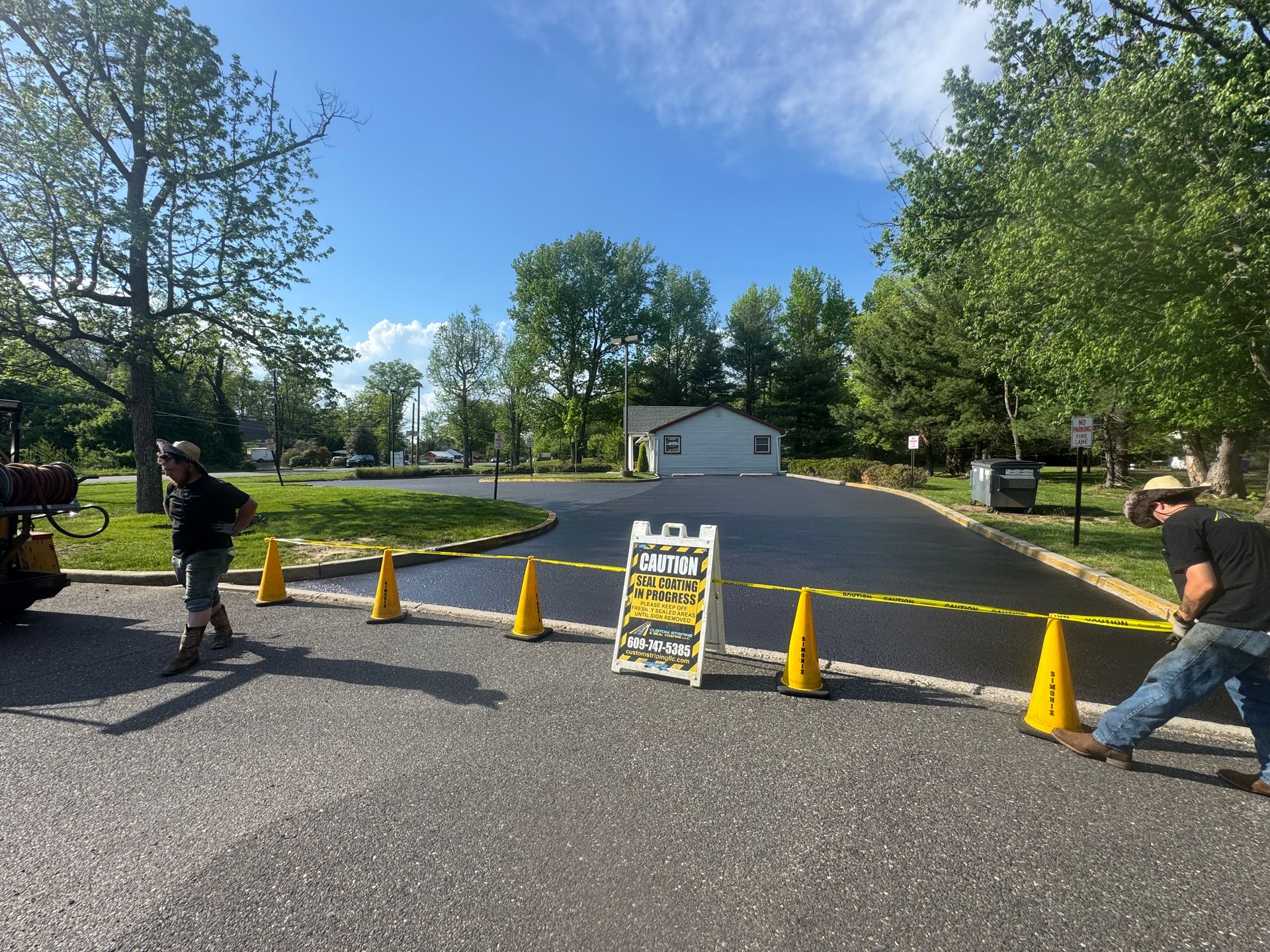 Workers roping off new asphalt driveway; cones, sign, and small building in background, sunny day.