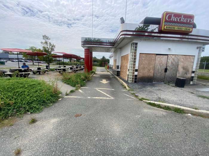 Abandoned Checkers restaurant with boarded-up windows, overgrown weeds, and outdoor seating area.