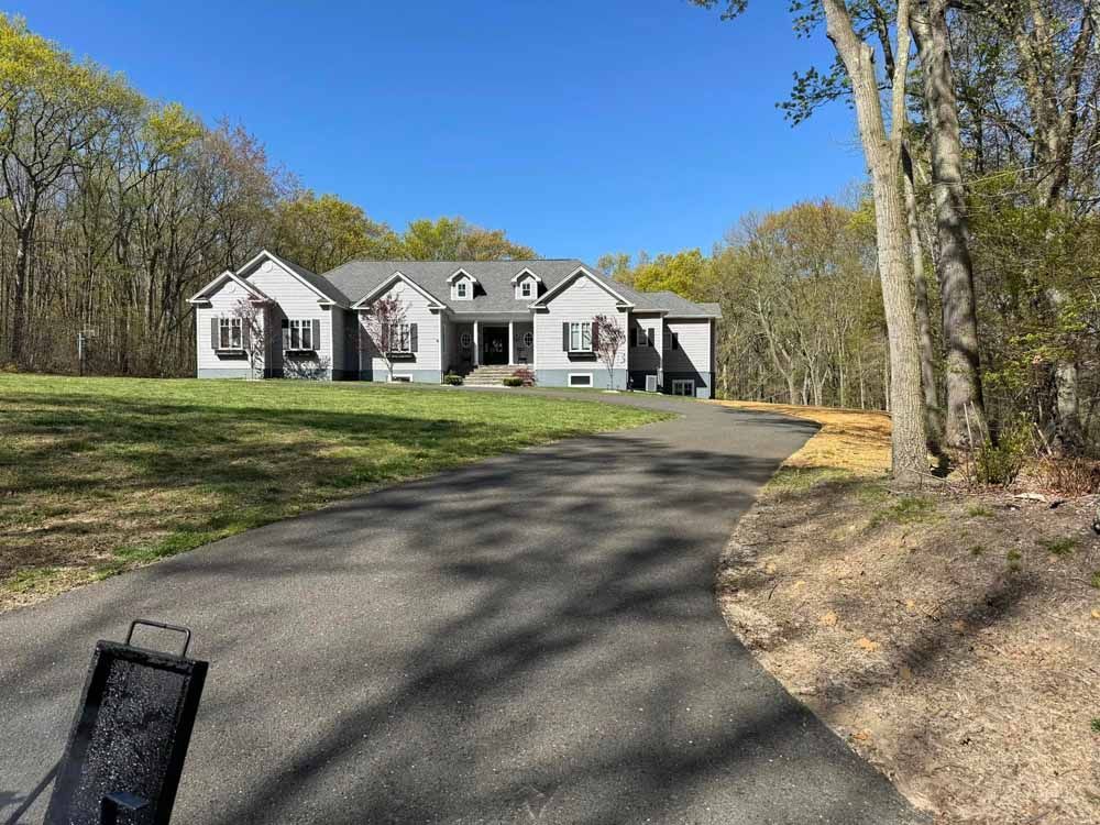 Gray house with long driveway on a sunny day surrounded by trees.