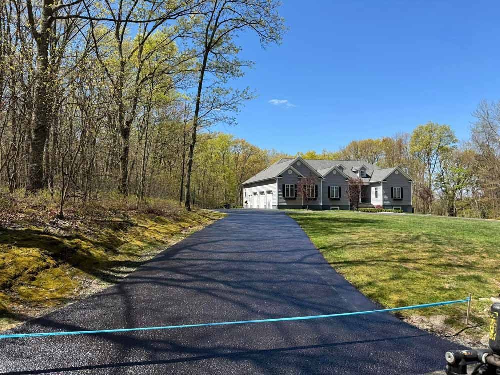 Asphalt driveway leads to a gray house with a three-car garage, on a grassy hill, with trees in the background.