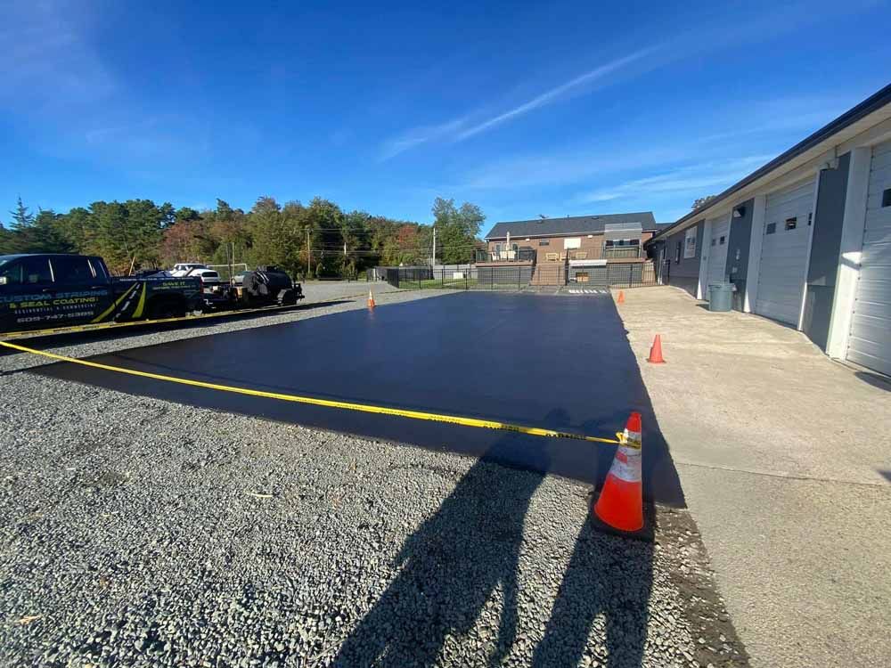 Newly paved asphalt parking lot, orange cones, caution tape, blue sky, and a building on the right.