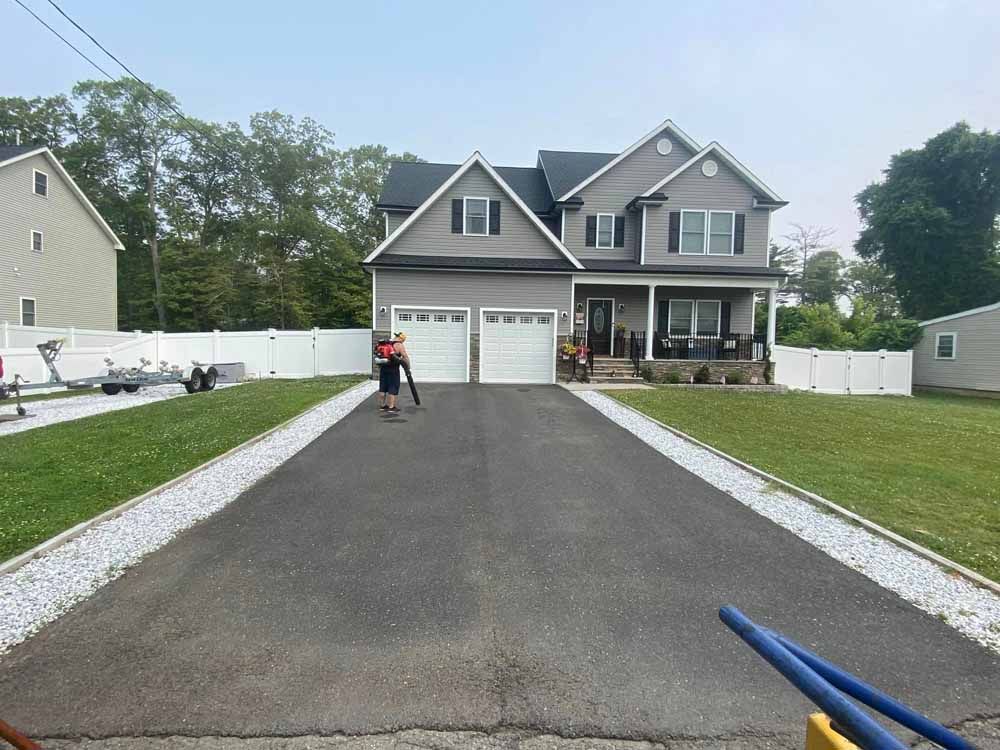 Person using a leaf blower on a black asphalt driveway in front of a gray two-story house.