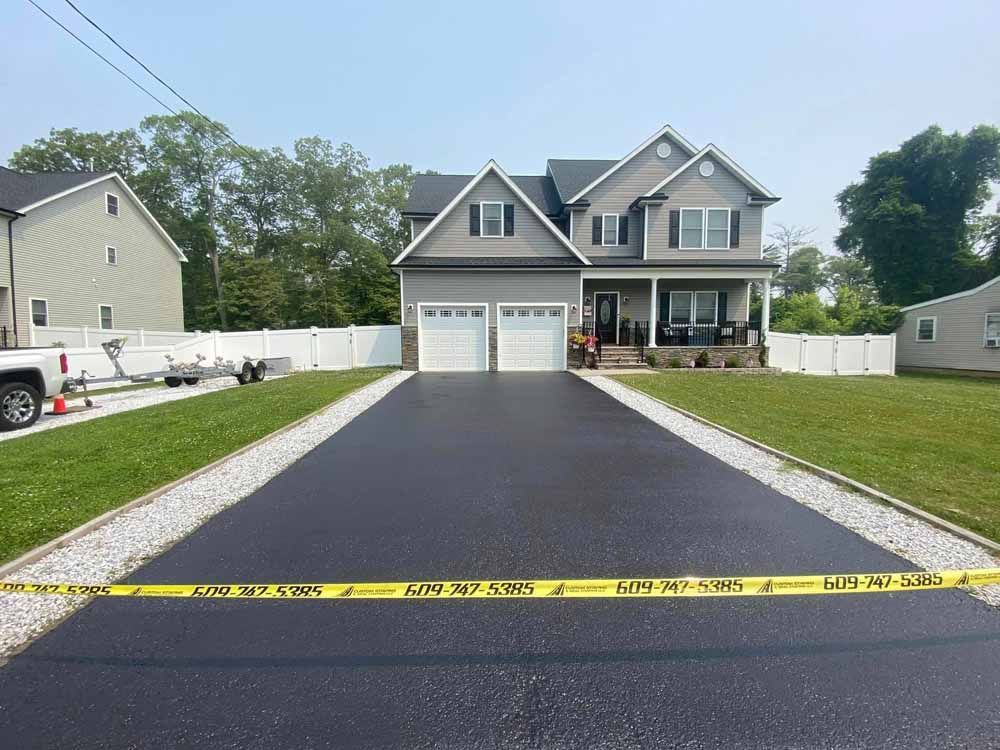 Newly paved asphalt driveway leading to a two-story gray house with a white fence on each side.