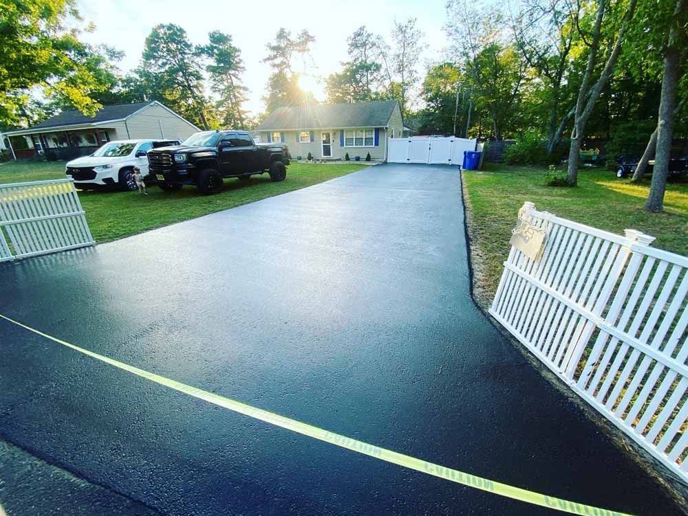 Freshly paved black driveway leading to a house with vehicles parked on the grass; sunny day.