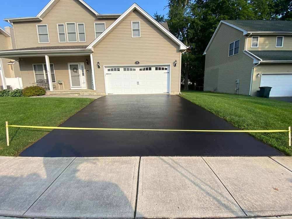 Newly sealed black driveway in front of a tan two-story house, bordered by green grass and a sidewalk.