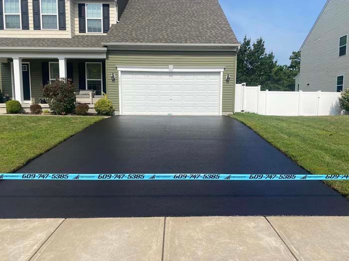 Freshly sealed black asphalt driveway in front of a house, bordered by green grass and a sidewalk.