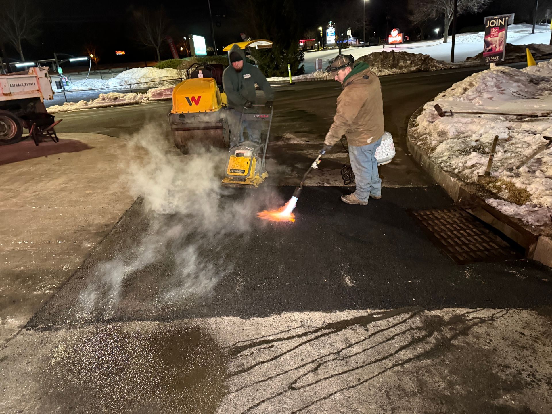 Workers use a snowblower and torch to clear ice from a snowy parking lot at night.