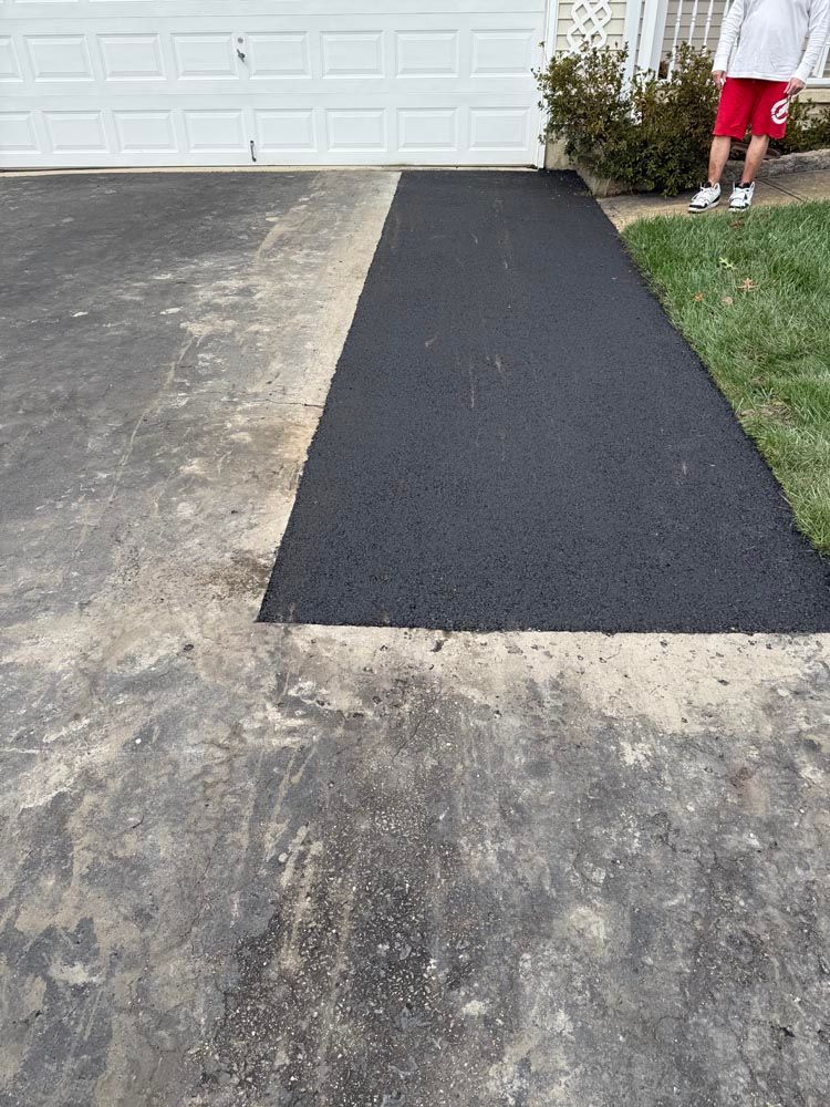 Driveway with a new black asphalt patch leading to a garage; part of a person in red shorts stands at right.