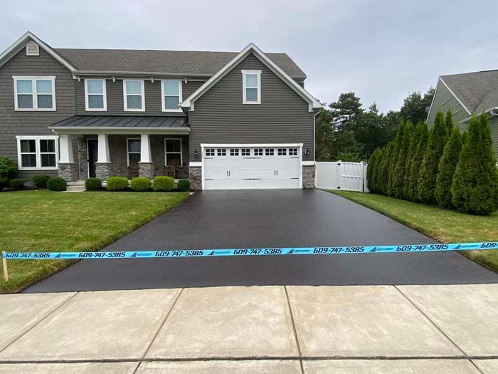 Newly paved asphalt driveway in front of a gray house with green grass and white garage door. Blue caution tape is across the driveway.