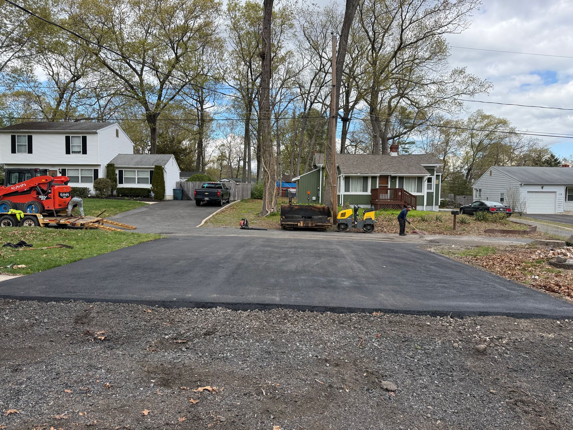 Freshly paved driveway in a suburban yard with houses, trees, and construction equipment nearby