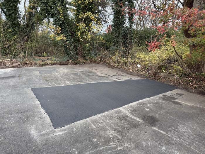 Fresh black asphalt patch on a worn concrete driveway beside trees and shrubs