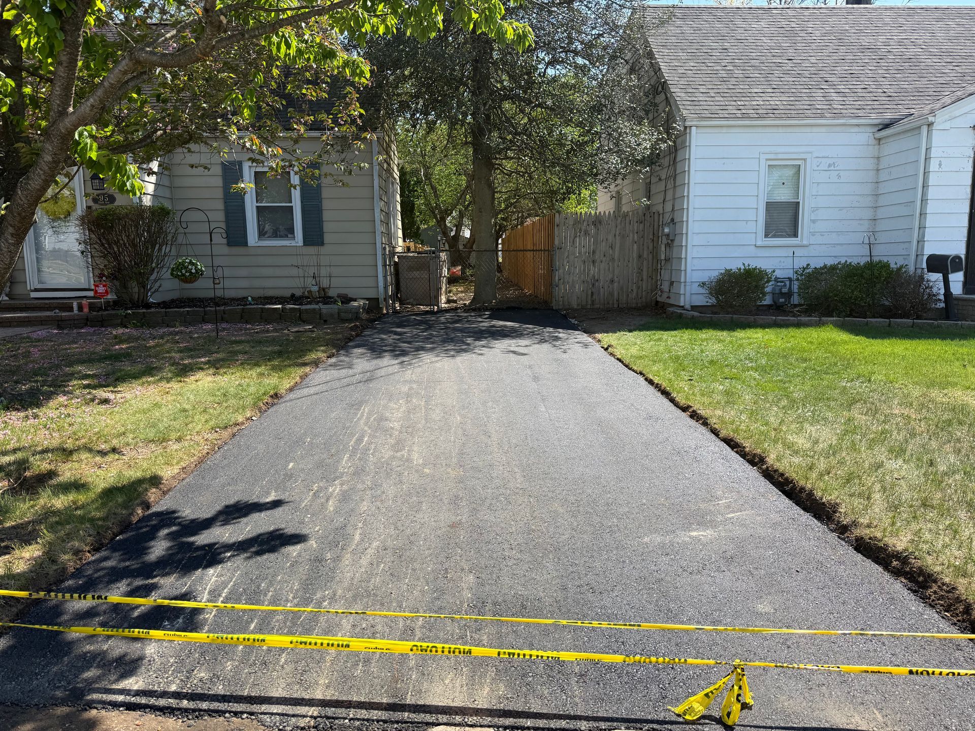 Black asphalt driveway leading to a house, bordered by grass and trees, with two yellow caution lines across it