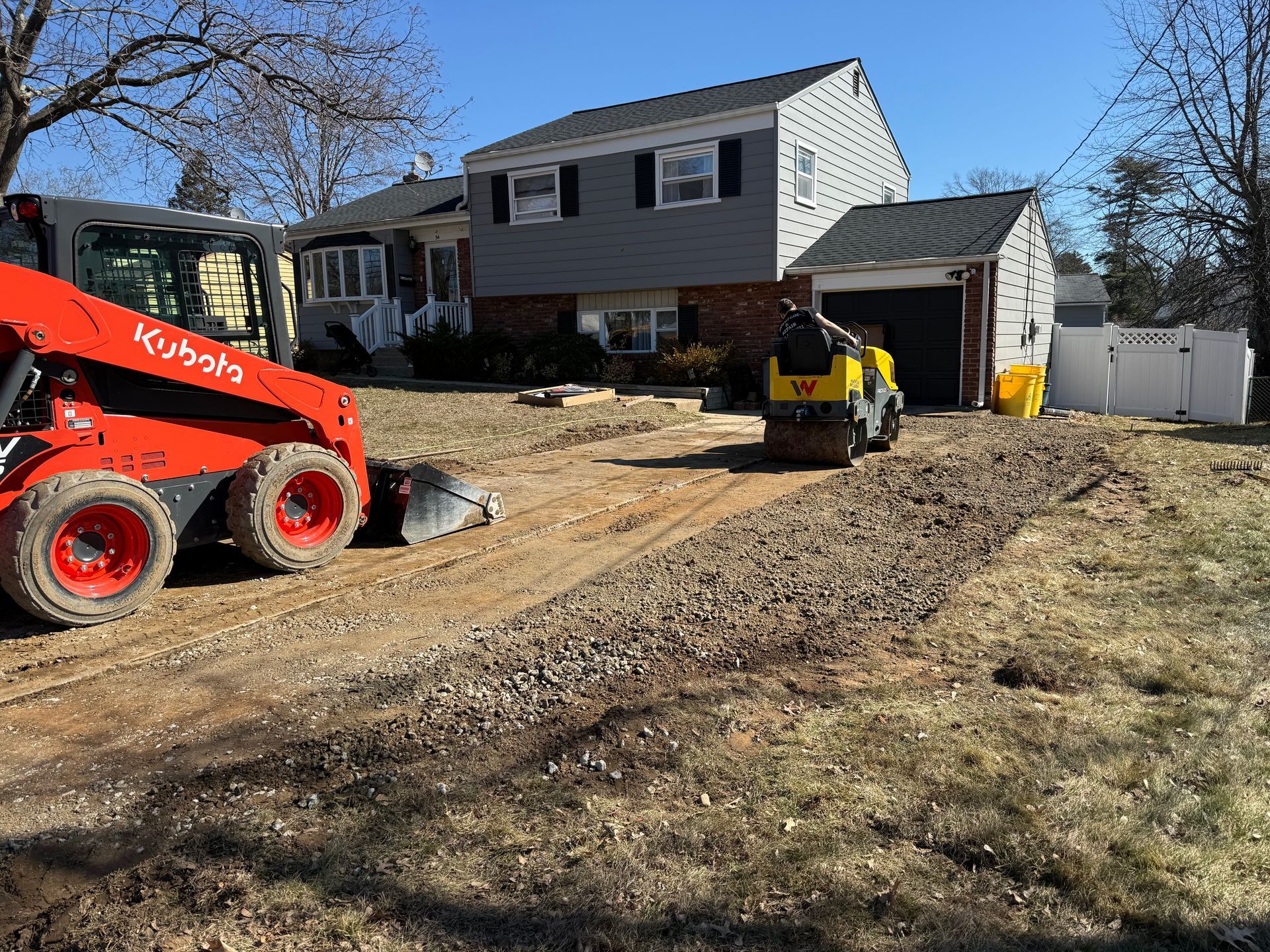 Workers with a skid steer and dump trailer grading a dirt driveway beside a suburban house