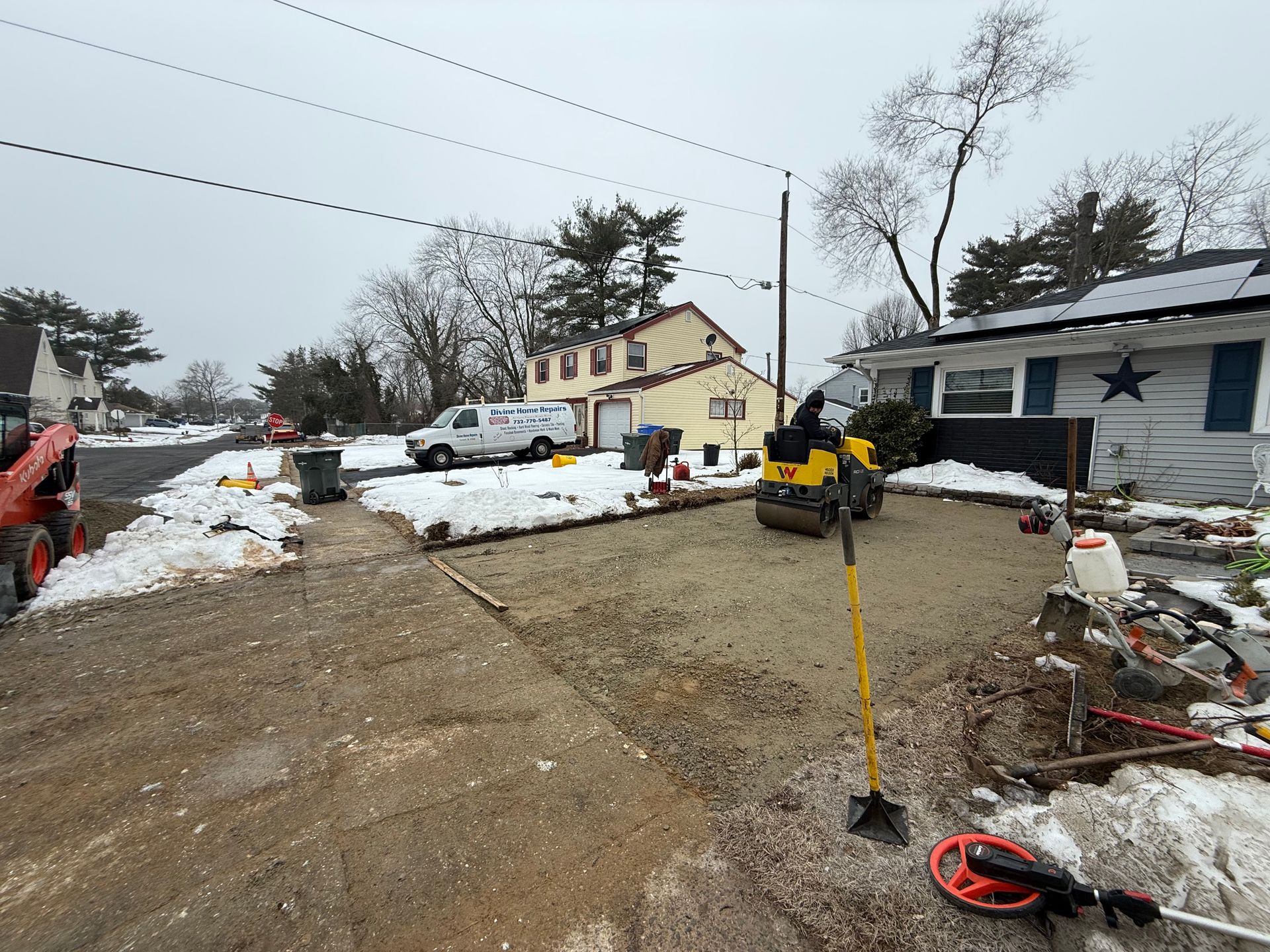 Snowy construction site with two workers, machinery, and houses under a cloudy sky