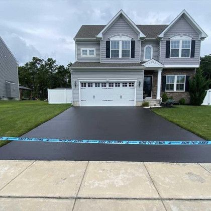 Two-story gray house with newly paved, black driveway, blue caution tape, green lawn, and sidewalk.