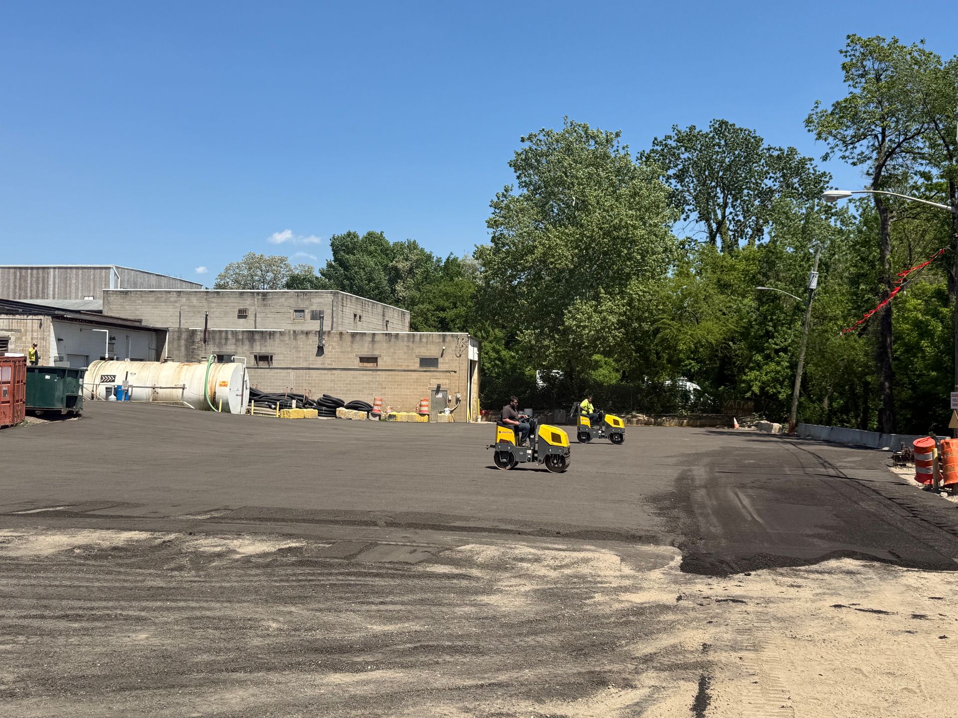 Wide construction yard with small yellow utility vehicles, a concrete building, and trees under a blue sky