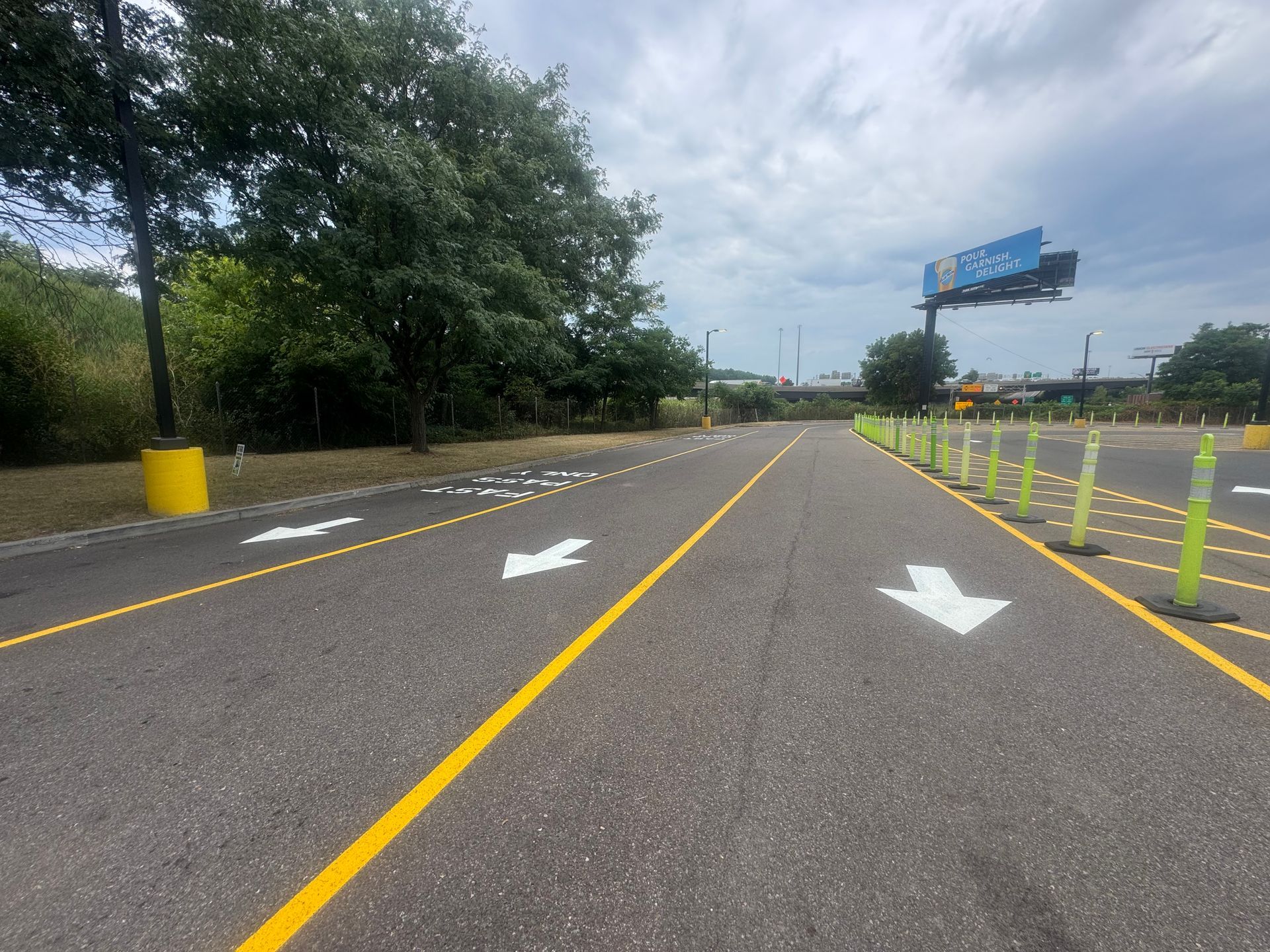 Asphalt road with yellow lines and white arrows directing traffic, yellow cones on the right, trees on the left.