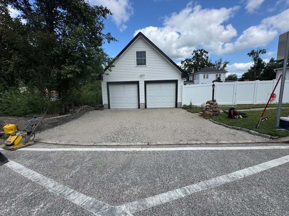 White garage with double doors at the end of a gravel driveway, bordered by trees and a white fence.