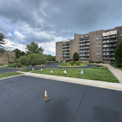 Large brick apartment building with balconies, cloudy sky, green landscaping, asphalt parking lot, traffic cone.