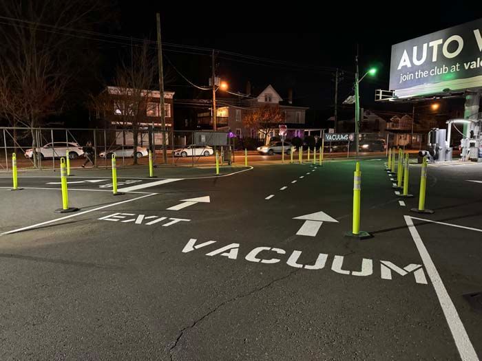 Asphalt parking lot at night. Yellow bollards delineate vacuum area and exit. Illuminated sign for auto wash in background.