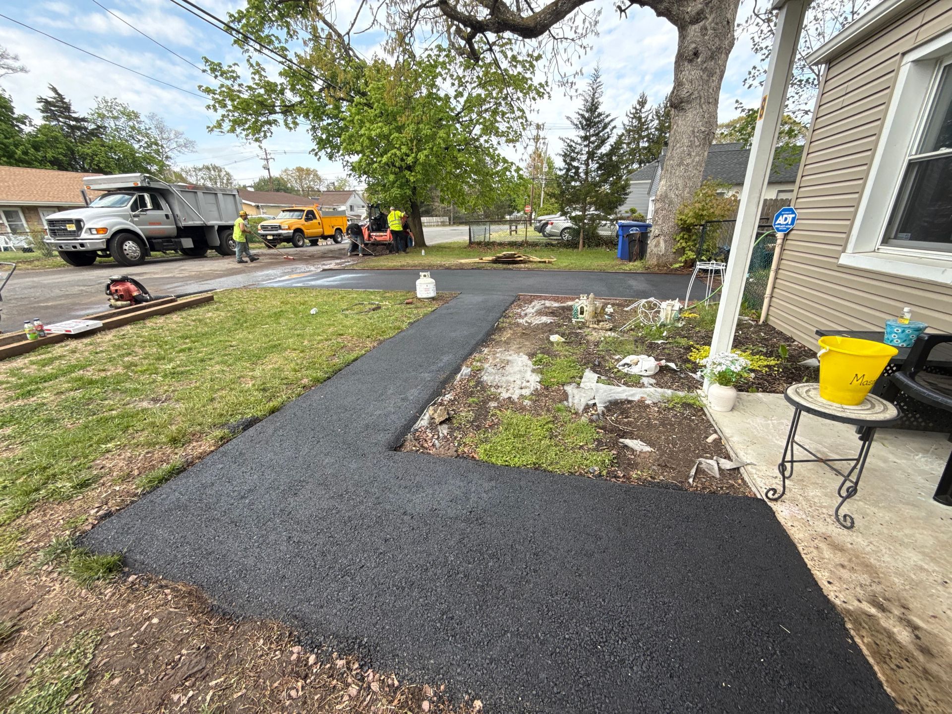 New black asphalt walkway beside a house, bordered by a garden bed and yard equipment.