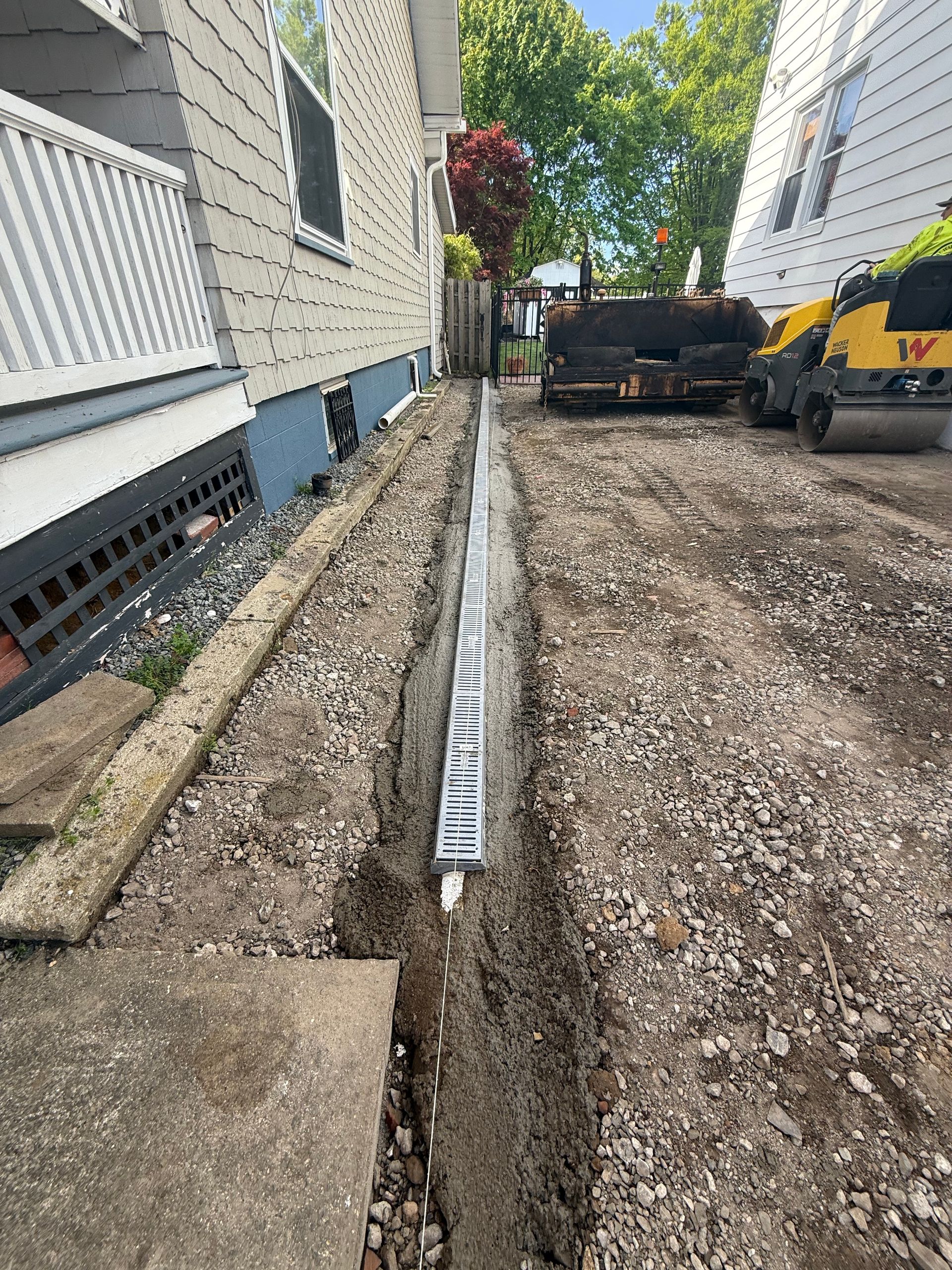 Narrow gravel trench beside houses with a long white drainage pipe and construction equipment in the background