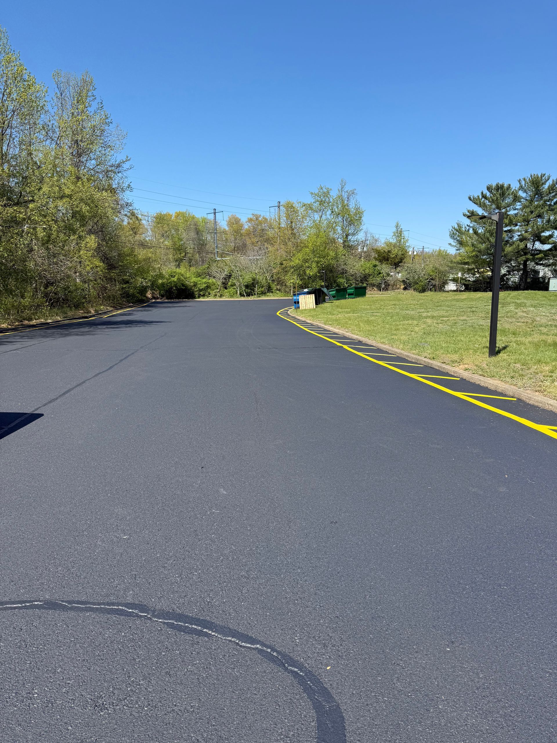 Curving paved road beside grass and trees under a clear blue sky, with yellow edge lines on the right