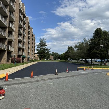 Newly paved asphalt in front of a brick apartment building, marked with traffic cones. Blue sky with clouds.