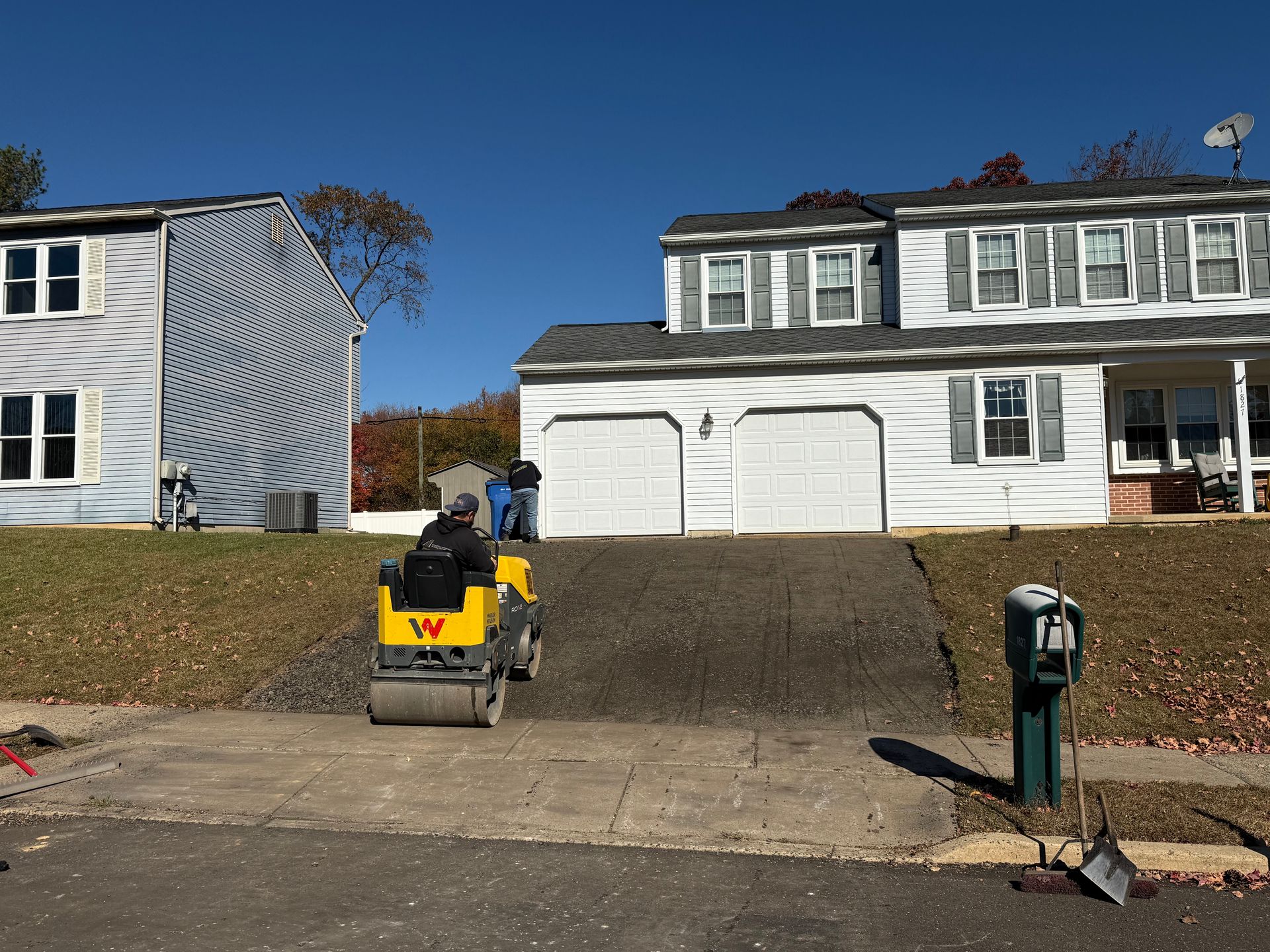 Person on yellow compact loader on a driveway in front of a white house under a clear blue sky