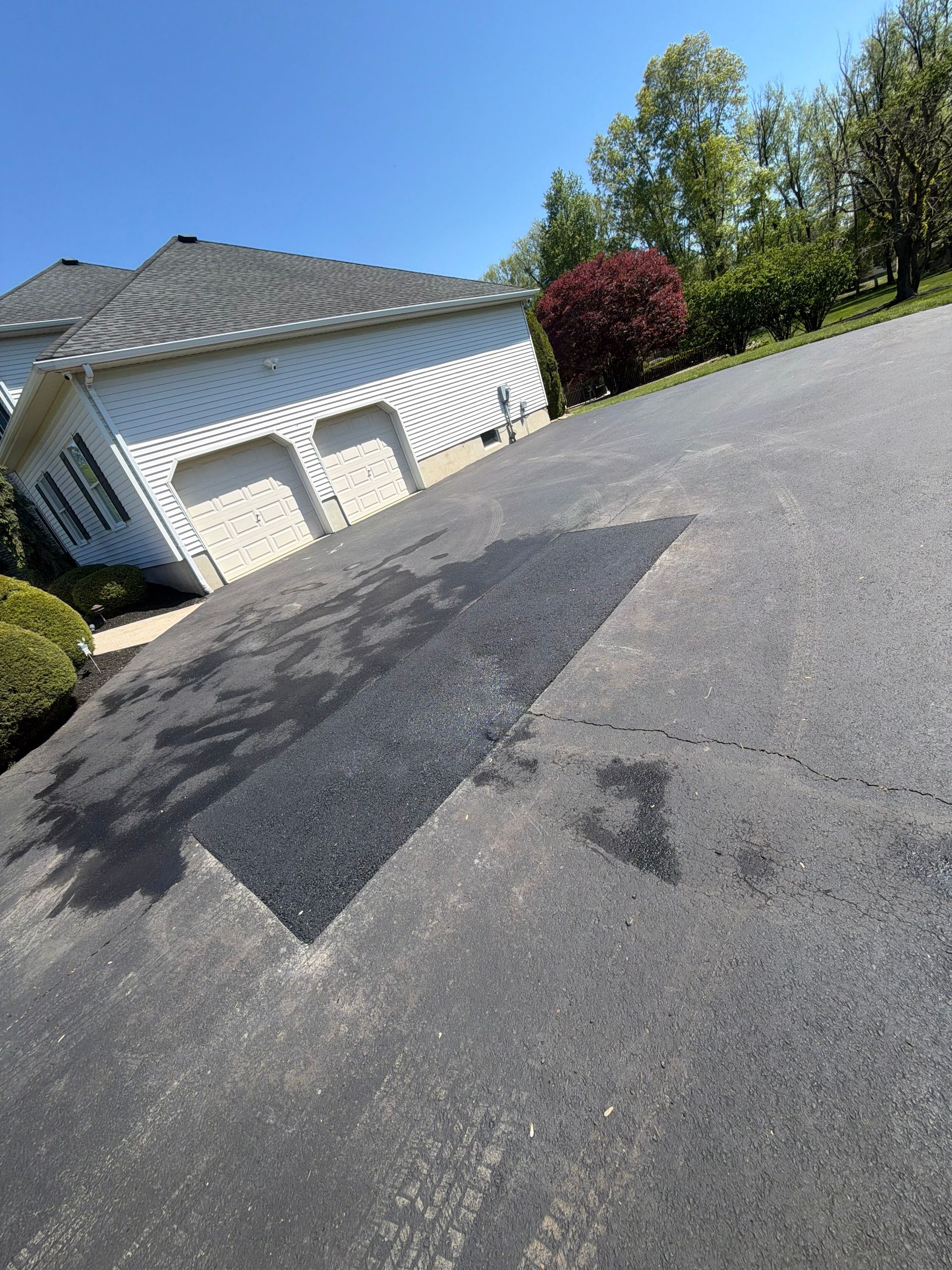 Tilted view of a suburban driveway with garage, fresh asphalt patch, trees, and blue sky
