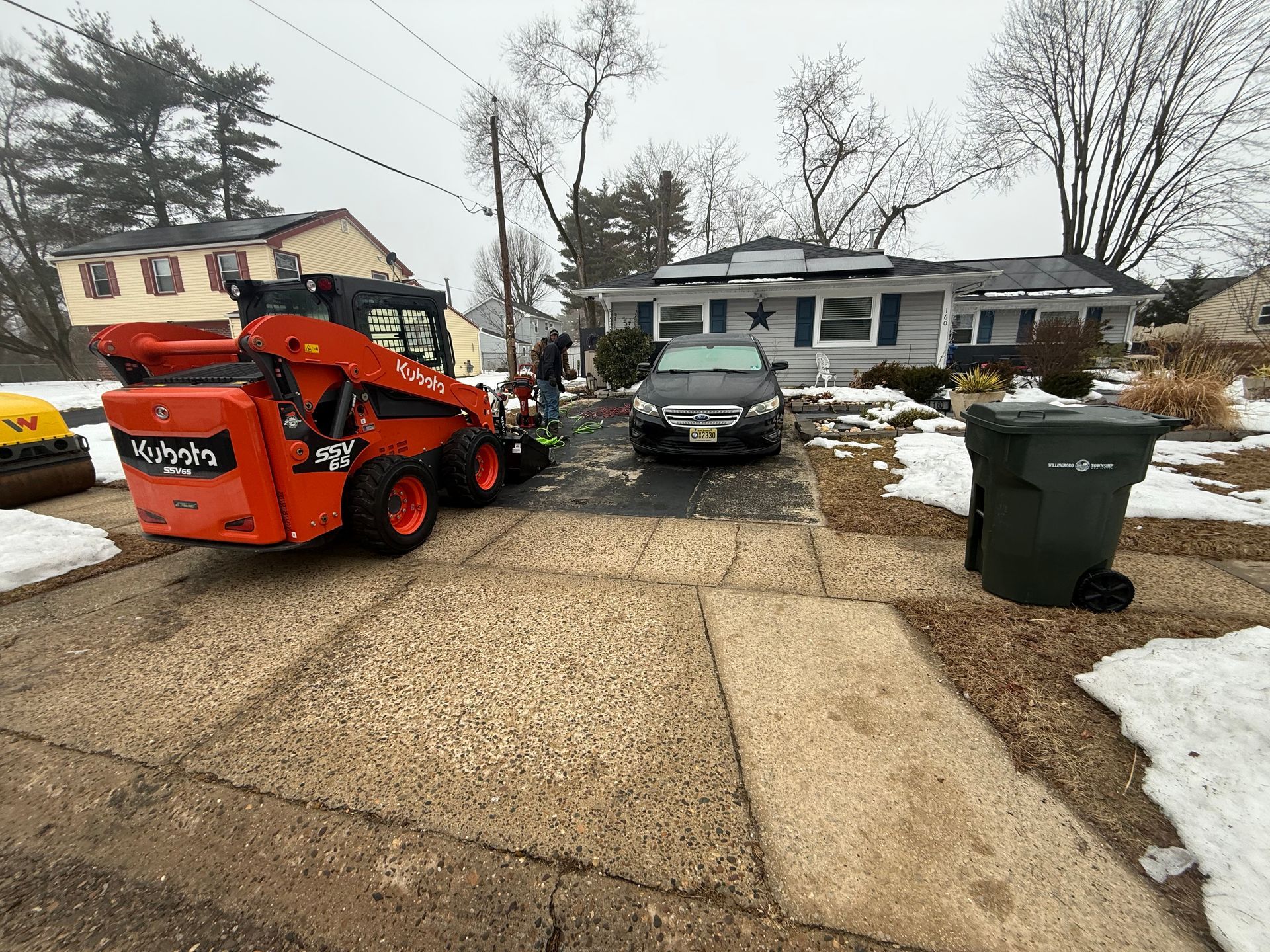 Orange Bobcat loader on snowy driveway with damaged black car in front of a house