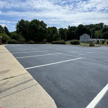 Newly paved asphalt parking lot with white painted lines, sunny day. Trees and building in background.