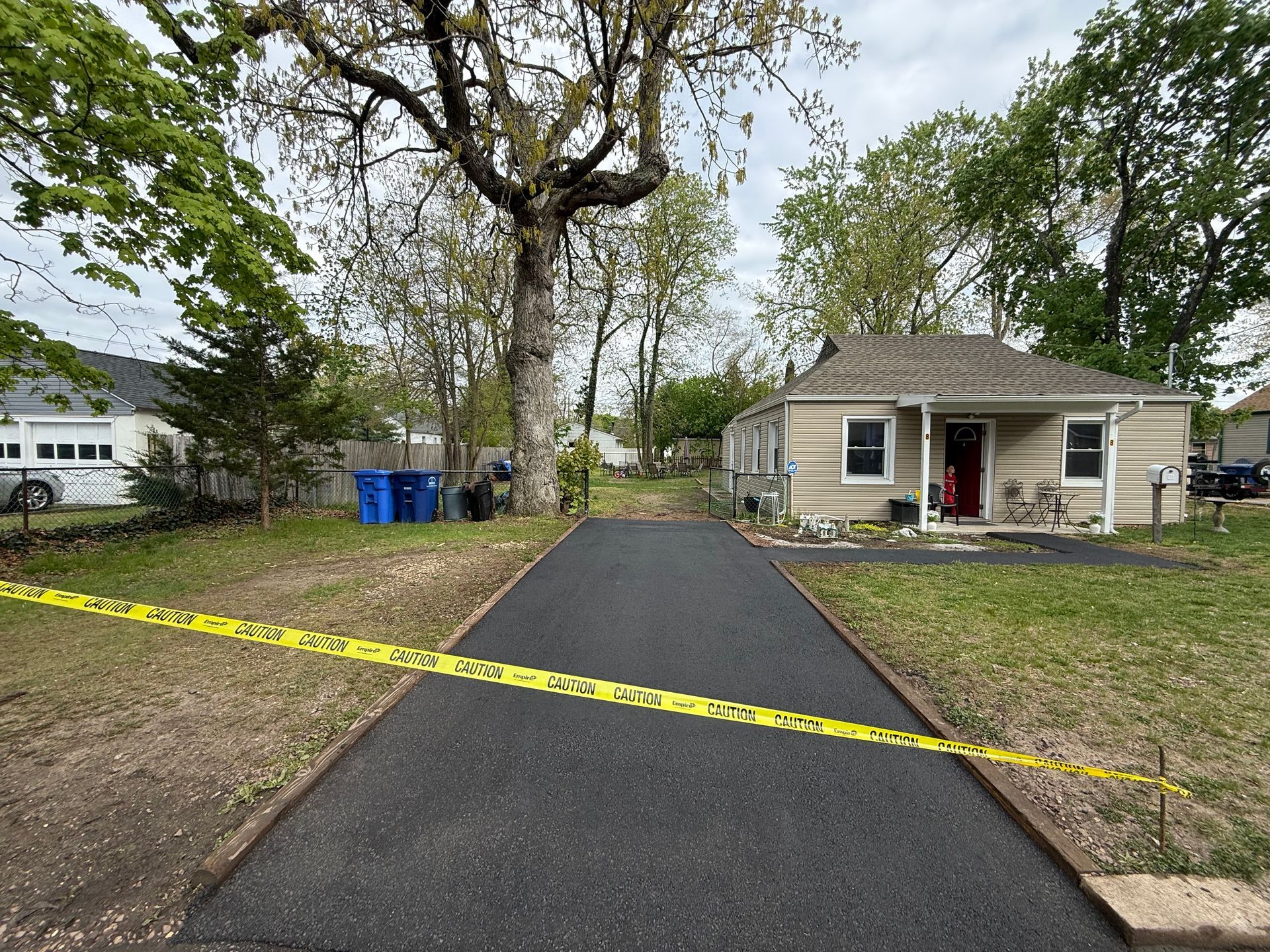 Yellow caution tape blocks a driveway leading to a small house and yard with large trees.