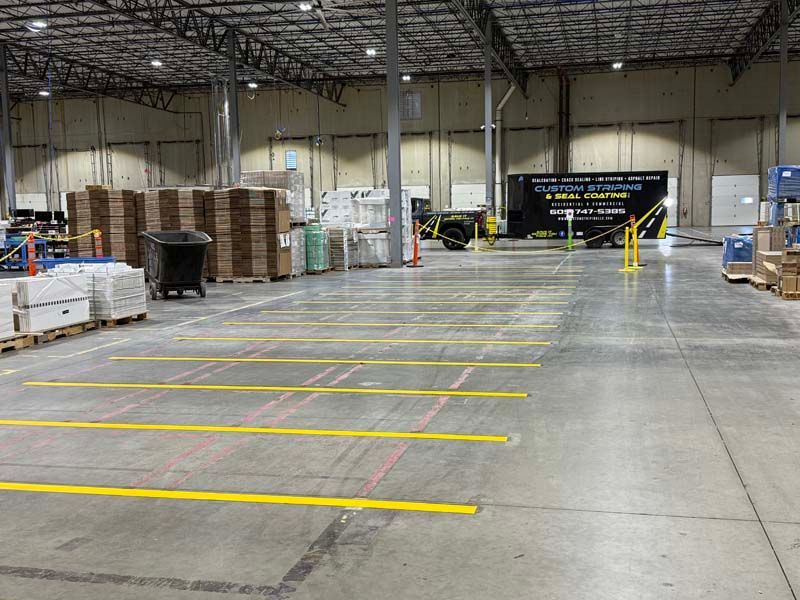 Warehouse interior with yellow floor lines. Pallets of boxes, trash can, and vehicle present.