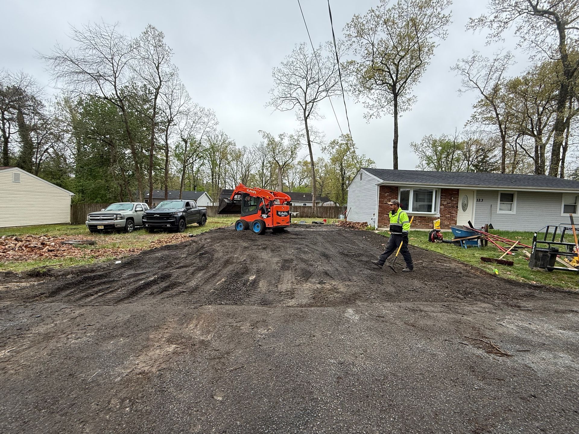 Construction site with muddy driveway, orange excavator, worker in neon vest, and house in background
